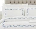 White bedding set with a light blue floral border pattern, styled on a bed with matching pillows and a tufted headboard.