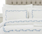White bedding set with a light blue floral border pattern, styled on a bed with matching pillows and a tufted headboard.