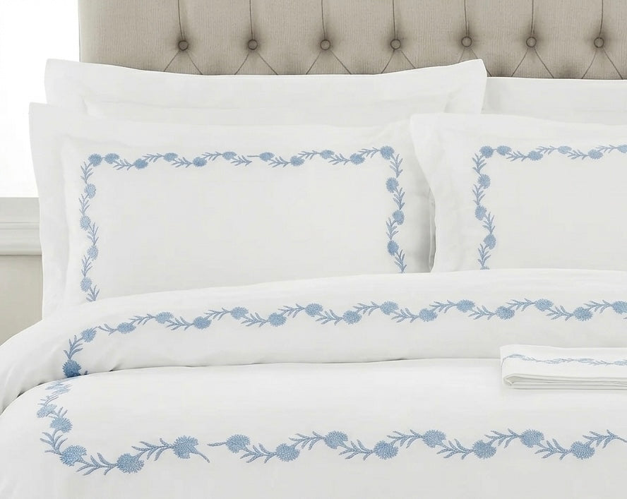 White bedding set with a light blue floral border pattern, styled on a bed with matching pillows and a tufted headboard.