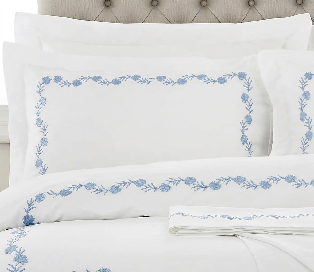 White bedding set with light blue floral border embroidery on the pillowcases and duvet cover, styled with a tufted headboard.