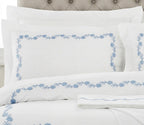 White bedding set with light blue floral border embroidery on the pillowcases and duvet cover, styled with a tufted headboard.