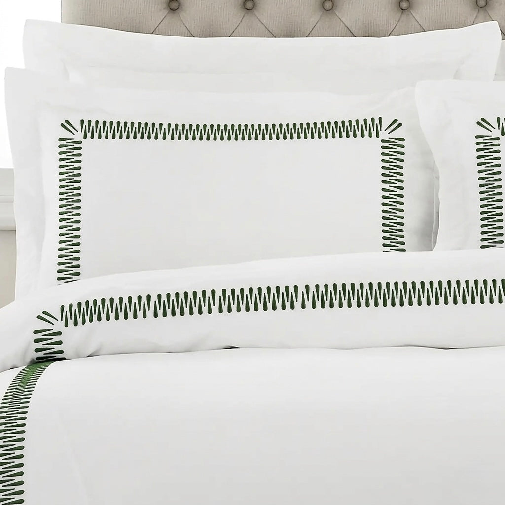 White bedding set with green zigzag border embroidery on the pillowcases and duvet cover, styled on a bed with a tufted headboard.